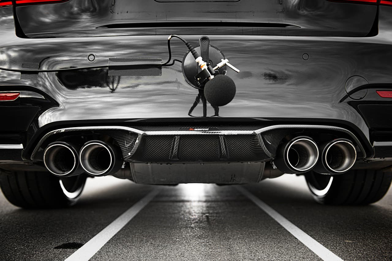 Four exhaust tips under a dark car, a sound-measuring microphone set up behind